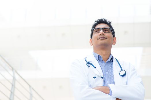 Portrait of a smiling Asian Indian male medical doctor in uniform standing in front of hospital looking away.
