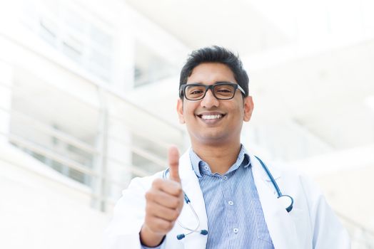 Portrait of a smiling Asian Indian male medical doctor in uniform thumb up, hospital building at background.