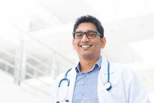 Portrait of a smiling Asian Indian male medical doctor in uniform, hospital building at background.