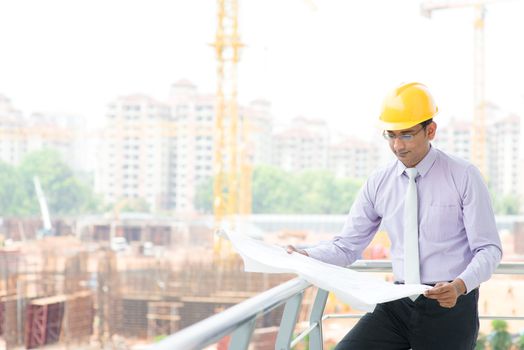 Portrait of a smiling Asian Indian male contractor engineer with hard hat reading blueprint at construction site.