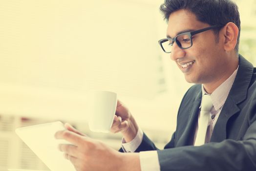 Young Asian Indian businessman using a tablet computer during office break at cafe, relaxing with a cup of coffee. India male business man,  in vintage retro style.