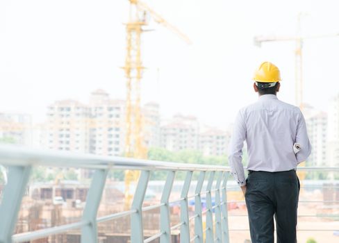 Rear view of a Asian Indian male contractor engineer with hard hat standing in front construction site, inspecting the progress.