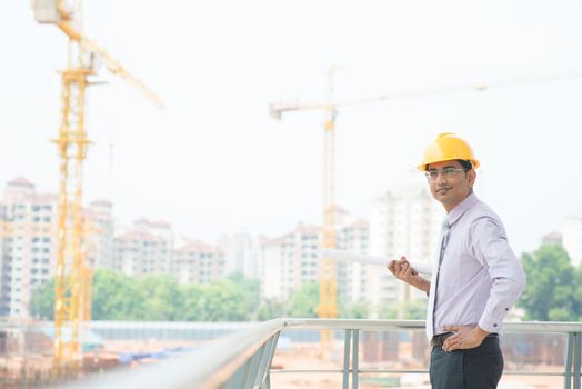Portrait of a smiling Asian Indian male contractor engineer with hard hat standing at construction site.
