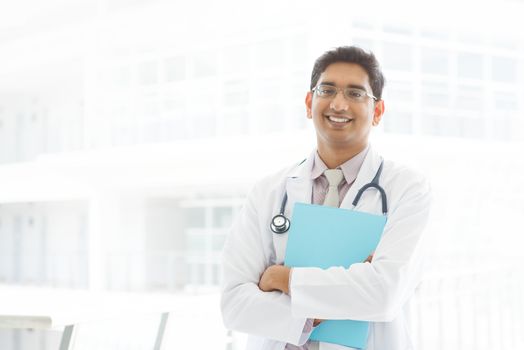 Portrait of a smiling Asian Indian male medical doctor standing inside hospital, holding file folder.