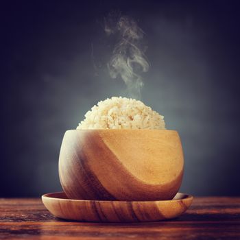 Cooked organic basmati brown rice in wooden bowl with hot steam smoke on dining table. Low light setting with retro revival style.