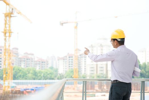 Portrait of a smiling Asian Indian male contractor engineer with hard hat pointing to construction site.