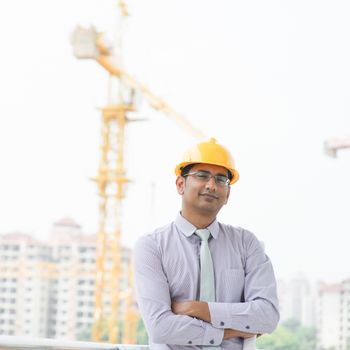 Portrait of a smiling Asian Indian male contractor engineer with hard hat standing in front construction site.