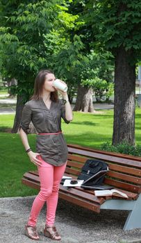 Young woman standing-up near a bench in a park and drinking a hot coffee during a break of her work on a laptop.
