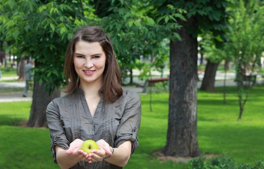 Young smiling woman offering a green apple outside in a park in summer.