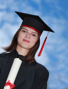 Portrait of a young woman in graduation gown against a cloudy sky.