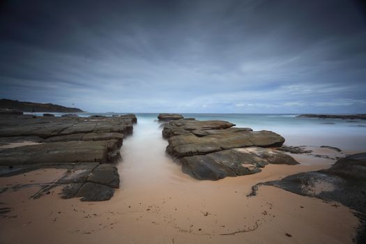 Stormy beach with foreground rocks at the edge of Soldiers point Norah Head NSW Australia.  Norah Head Lighthouse in distance. Long exposure