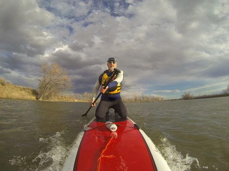 mature male paddler in wetsuit and life jacket enjoying workout on a stand up paddleboard (SUP), windy lake in one of nature areas in Fort Collins, Colorado,  distorted wide angle view