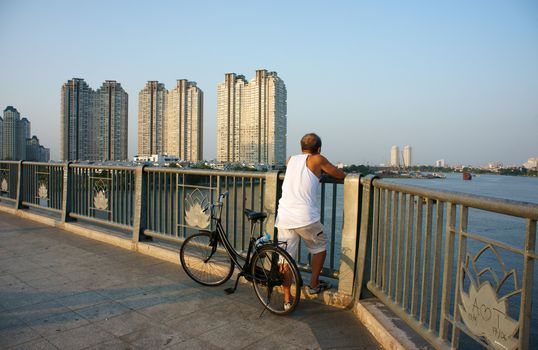HO CHI MINH, VIET NAM- MAR 12: Healthy lifestyle of citizen at evening, ride bicycle to do exercise, stand on handrail of bridge to relaxed, enjoy a view, building at riverside, Vietnam, Mar 12, 2014