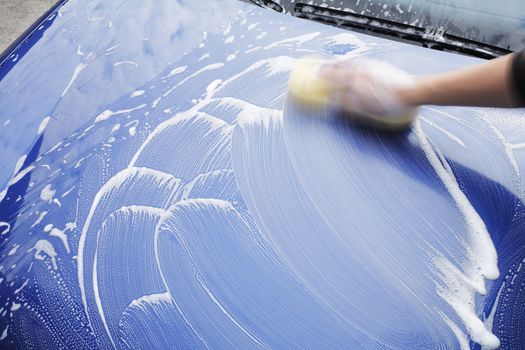 A motion blurred image of a hand washing the hood of a blue car with a sponge