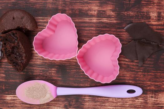 Baking delicious muffins. Chocolate baking mixture, fresh muffins and pink heart shaped baking forms on brown wooden background, top view.