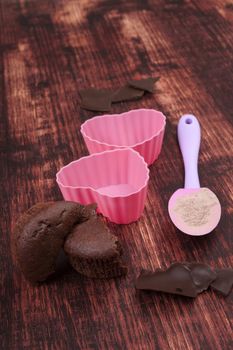 Chocolate muffin, chocolate bar, baking form and baking mixtures on brown wooden background.