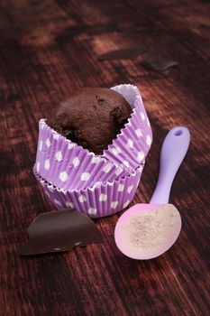 Chocolate cupcake in purple dotted paper baking form, chocolate bar and baking mixture on wooden background. Baking cupcakes rustic style.