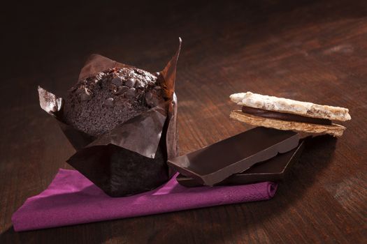 Delicious chocolate muffin with chocolate bar and cookie on dark wooden background. Sweet luxurious dessert eating.
