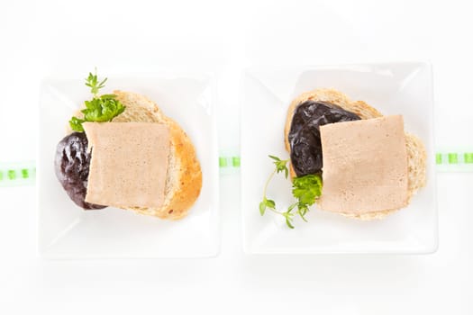 Two white bread pieces in white square bowls with delicious pate, dried plum and fresh herbs.