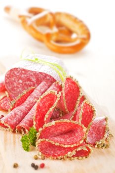 Salami slices on wooden board with fresh herbs and pepper corns. Pastry in background.