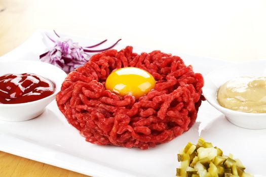 Steak tartare with yolk on white plate with onions, pickles, mustard and ketchup. Toast in background. 