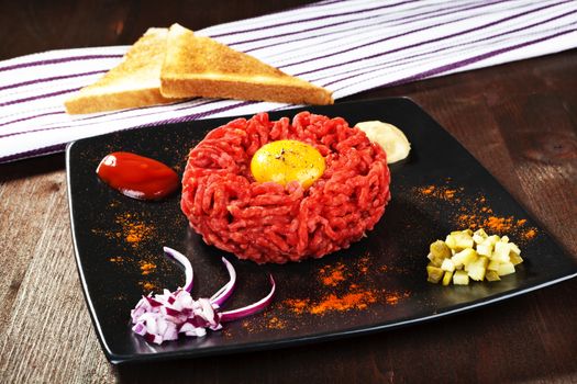 Steak tartare with yolk on black plate. With chopped onion, pickle, ketchup, mustard and toast bread. Gourmet dining.