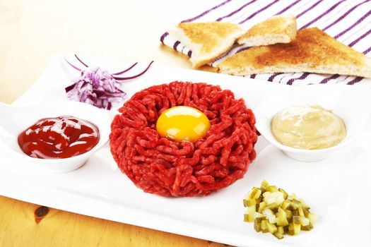 Luxurious steak tartare with yolk on white plate with ketchup, mustard, onion and pickle. Toast bread in background. Gourmet dining.