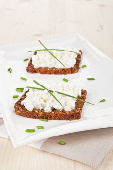 Delicious dark bread slices with cottage cheese and fresh herbs on white plate on wooden background.