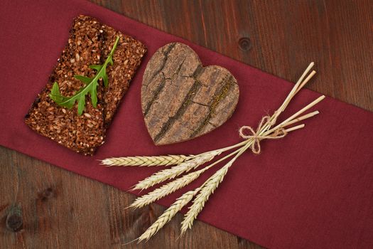 Black bread slice with wooden heart shape and bunch of wheat on dark wooden background. I love healthy food.
