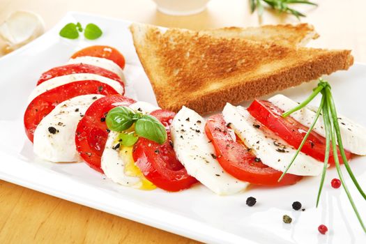 Fresh tomato and mozzarella slices arranged on white plate with toast bread and fresh basil leaves.