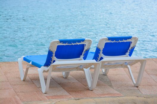 Two empty blue lounge chairs by the ocean.