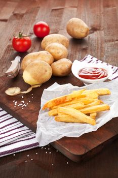 French fries on wooden board with potatoes and tomatoes in background. Country style.