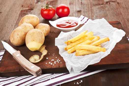 French fries on wooden board. Potatoes, tomatoes and ketchup in background.