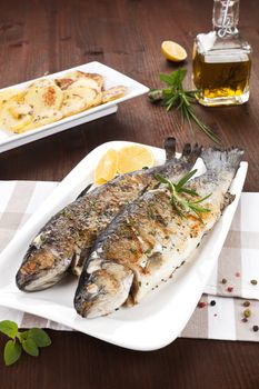 Two grilled trouts on white plate with lemon pieces, potatoes and olive oil on kitchen towel on wooden table.