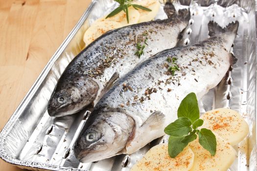 Two fish on grilling tray with potatoes and herbs prepared for grilling. Garlic and herbs in background.