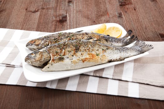 Grilled trout on white plate with lemon pieces on kitchen towel on wooden table.