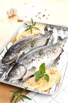 Two fish on grilling plate with potatoes and herbs prepared for grilling. Garlic and herbs in background.