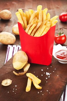 French fries in fast food paper bag. Potatoes, tomatoes and ketchup in background. Country style.