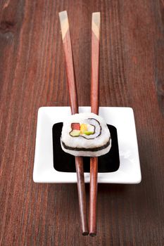 Maki sushi on chopsticks on soy sauce in white bowl on dark wooden background.