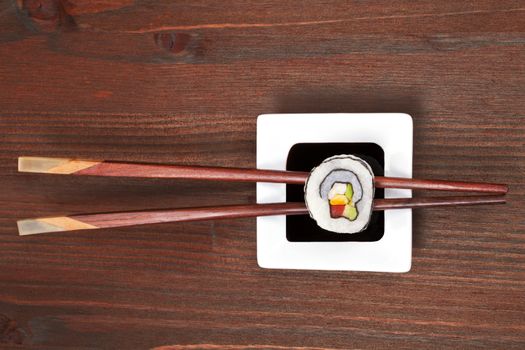 Maki sushi on chopsticks on soy sauce in white bowl on dark wooden background.