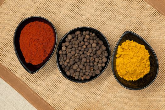 Three black bowls with different spices on brown natural background. Pepper corns, paprika and curry.