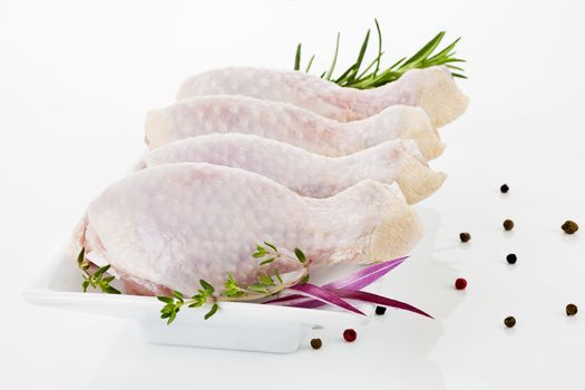 Four raw chicken legs on white plate decorated with rosemary, pepper corns and onion.