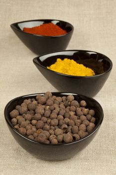 Three black bowls with different spices on brown natural textile background. Pepper corns, paprika and curry.