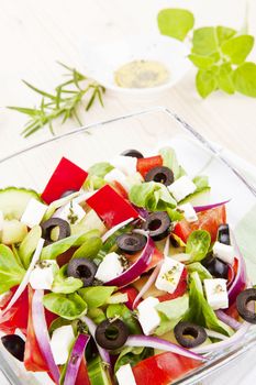 Greek salad in bowl close up, olive oil dressing and fresh herbs in background.