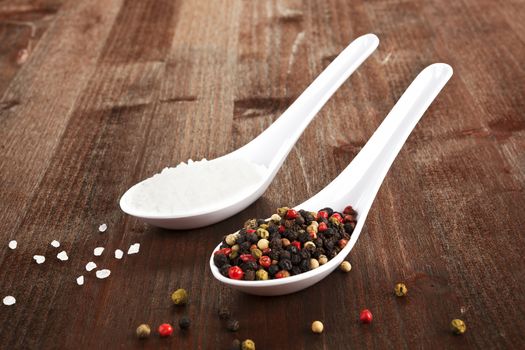 Sal crystals and colorful pepper corns on white spoons on dark wooden background.