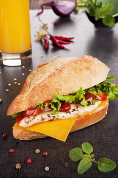 Sandwich with grilled chicken, vegetables and cheese. Pepper corns, chili peppers, fresh herbs and orange juice in background.