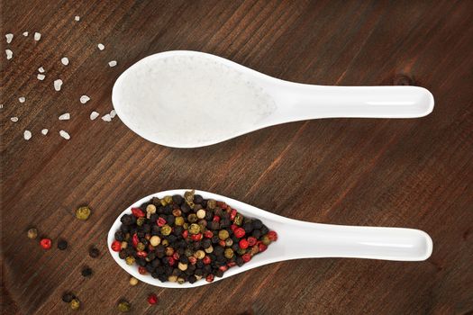 Salt crystal and pepper corns on white spoon on dark wooden background.