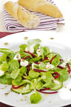 Fresh salad leaves with cucumber pieces, beet and goat cheese with white bread in background.