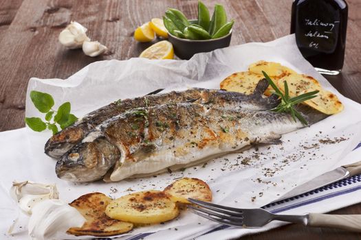 Two grilled trouts on white plate with lemon pieces, potatoes and balsamico on kitchen towel on wooden table.