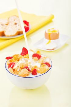 Pouring milk into a bowl with corn flakes and strawberries. Delicious breakfast. Egg and pastry in background.